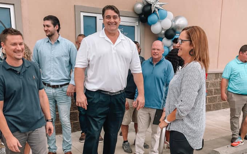 A group of people, both men and women, stand outside a building decorated with a balloon arch. They are casually dressed and smiling, appearing to engage in friendly conversation.