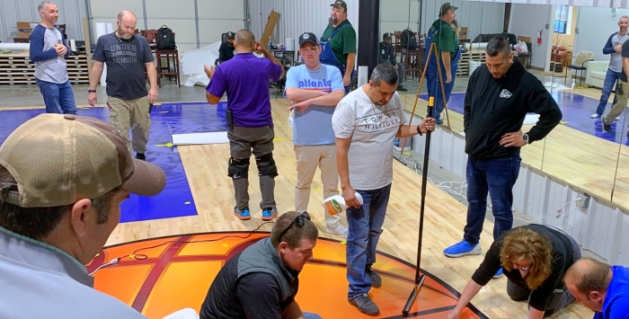 A group of people in casual attire are gathered in a large indoor space with mirrors and a wooden floor. Some are installing or laying down materials on a section of the floor, resembling a basketball court. Tools and equipment are visible around them.